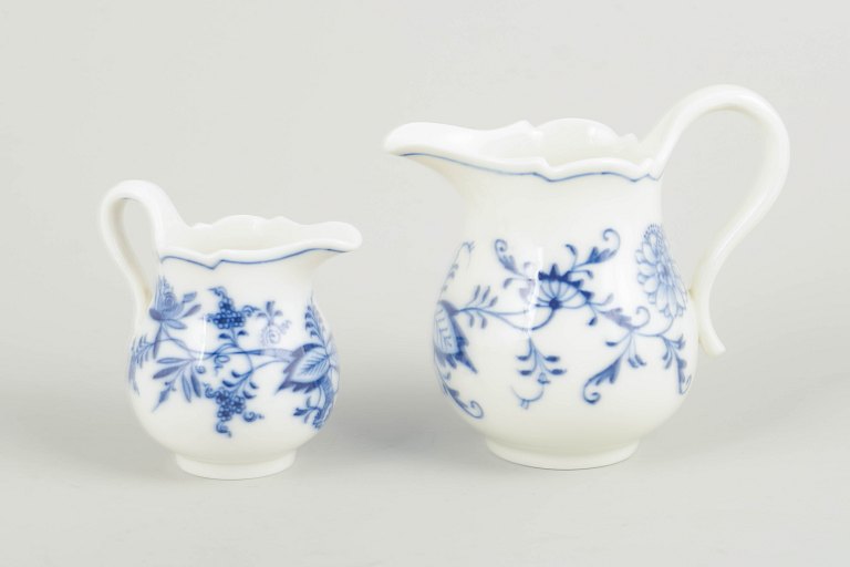 MeissenTo flødekander, “løgmønstret”, ca. 1900.To flødekander i Meissens klassiske “Zwiebelmuster”-dekoration, håndmalet i koboltblå på hvidt porcelæn. Organiske svungne korpusformer med buet hank og bølget kant. Oprindelse: Tyskland, Meissen.Designer: Meissen Porzellanmanufaktur.Materiale: Håndmalet porcelæn.Dimensioner: H. 8–11 cm, D. 8–12 cm inkl. hank.Periode: Ca. 1900.Stand: Mindre brugsspor; én kande i 1. sortering, én i 3. sortering.Bemærkning: Stemplet med Meissens korslagte sværd, det ene annulleret med to indridsede streger (3. sortering).Bio: Meissen Porzellanmanufaktur, grundlagt i 1710, er Europas ældste porcelænsfabrik og kendt for sin banebrydende udvikling af hårdt porcelæn. “Zwiebelmuster”, introduceret i 1730’erne, er et af manufakturens mest ikoniske og vedvarende mønstre, malet i koboltblå underglasur. Mønsteret er blevet produceret i flere århundreder og er i dag et højt værdsat samlerområde.